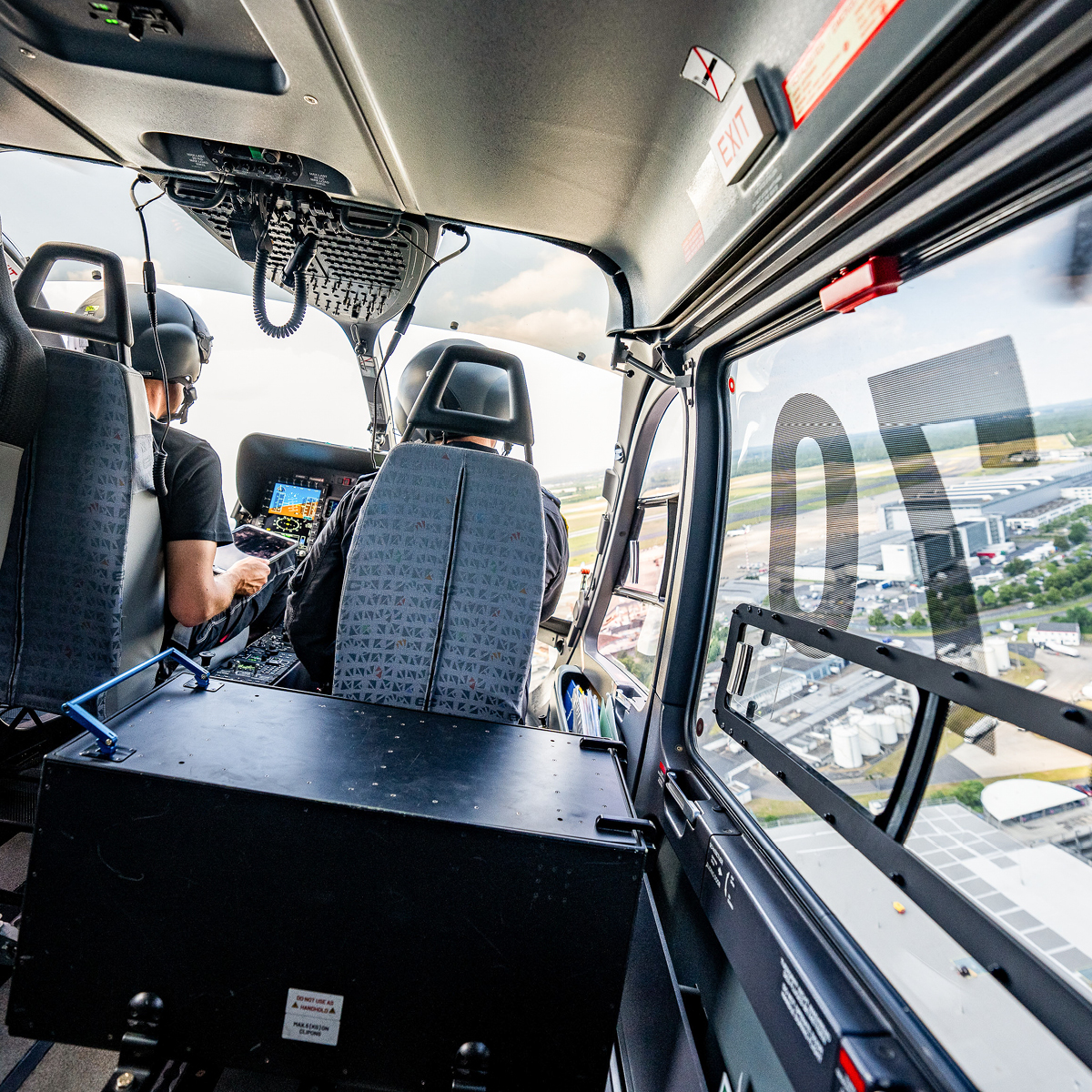 Das Foto wurde im hinteren Teil eines fliegenden Hubschraubers aufgenommen. Zu sehen sind Pilot und Copilot von hinten. Außerdem sieht man durch die Scheibe ein Industriegebiet.