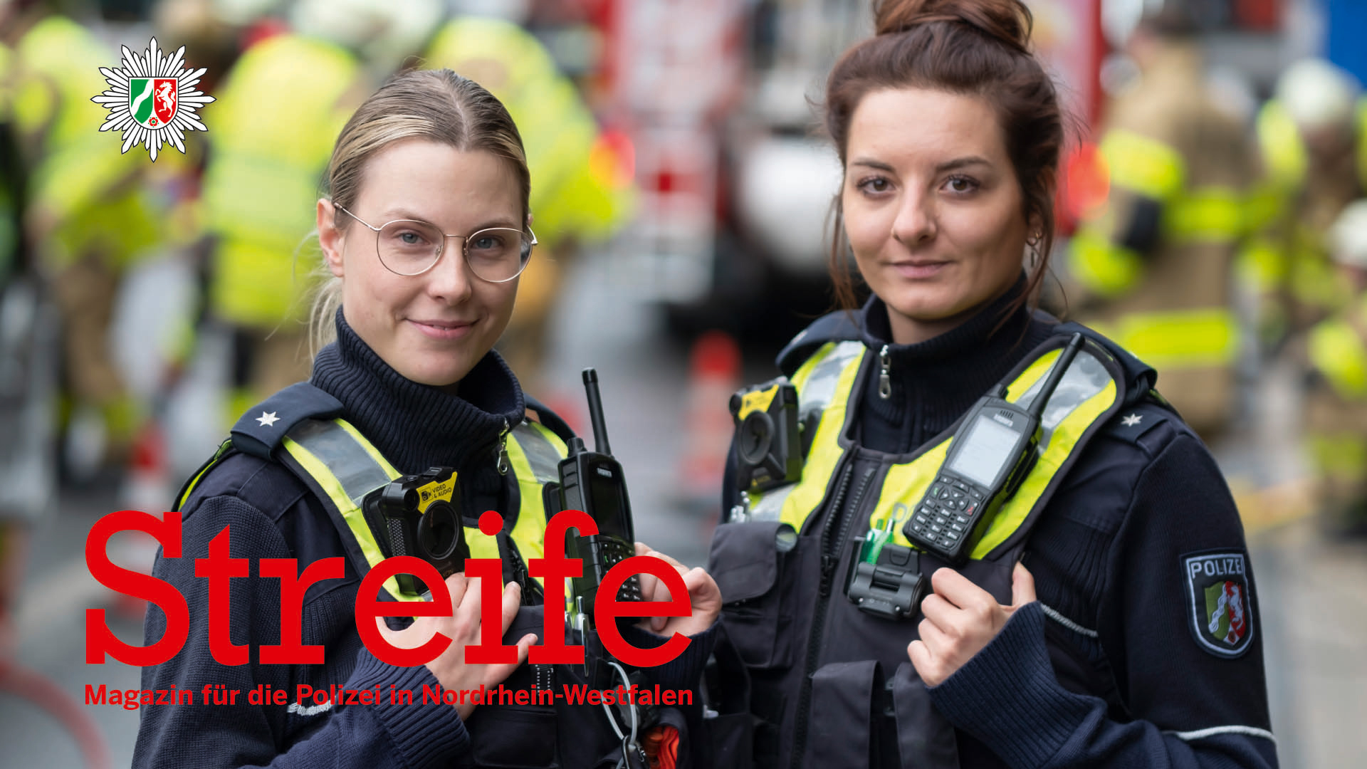 Im Vordergrund sind die Polizistinnen Paula Petzschke und Jennifer Krystkiewicz zu sehen. Sie tragen Uniform und haben die Hände an ihrer Weste eingehakt. Beide schauen in die Kamera. Im Hintergrund sind Rettungskräfte zu sehen.