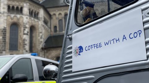 Logo der Aktion "Coffee with a Cop" auf einer Seitentür. Das Logo zeigt den Polizeistern und eine Tasse Kaffee in einem blauen Kreis sowie den Schriftzug Coffee with a Cop. Im Hintergrund ein Streifenwagen der Polizei und die Bonner Münsterkirche