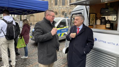 Innenminister Herbert Reul und Polizeipräsident Frank Hoever stehen vor dem Infostand zu Coffee with a Cop. Im Hintergrund weitere Personen, ein Streifenwagen der Polizei und die Bonner Münsterkirche.