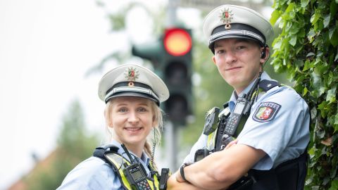 Uniformed policewoman on the left, uniformed police officer on the right, both wearing a white police cap on their heads, a blue protective vest, radio, holstered weapon and pepper spray. A red traffic light and a stop sign can be seen blurred in the background.